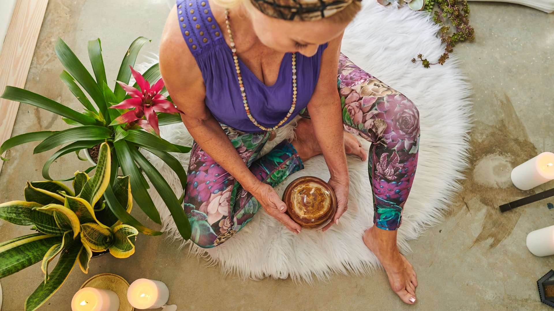 Jennifer seated cross-legged on a sheepskin holding a singing bowl, surrounded by plants and candles